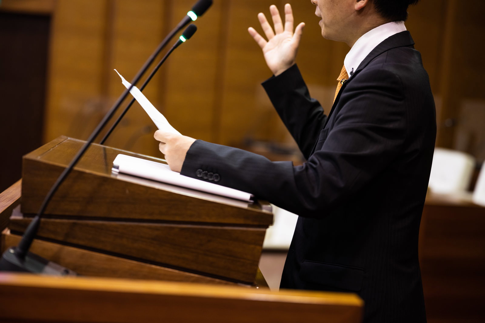 市議会の演壇で熱く語る黒いスーツの議員。手に書類を持ちながら説明している