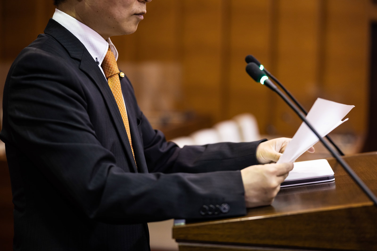 黒いスーツと橙色ネクタイの議員が原稿を手に議場のマイク前で答弁する