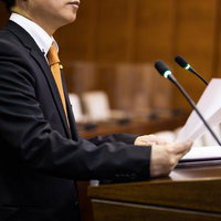 演壇で資料を手にするスーツ姿の男性議員の演説の写真