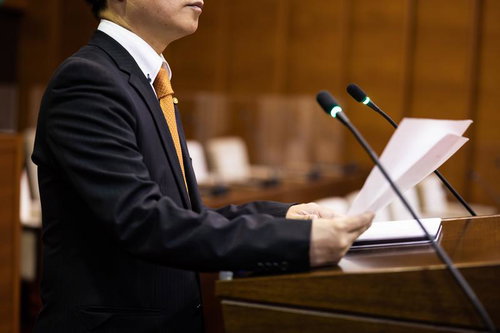 演壇で資料を手にするスーツ姿の男性議員の演説