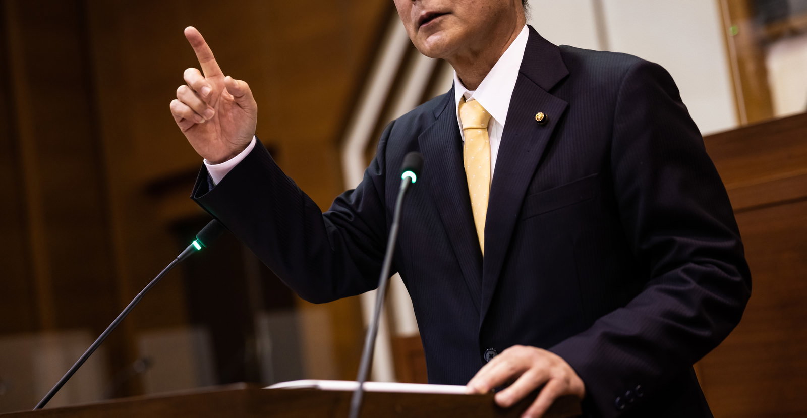 市議会の議場で手を挙げて発言する紺色の背広姿の議員
