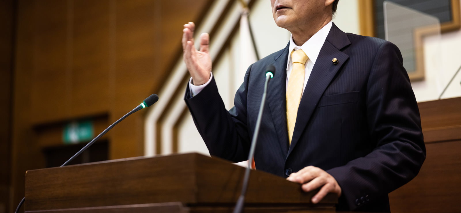 市議会の演台でマイクを前に片手を上げながら質問する市議会議員