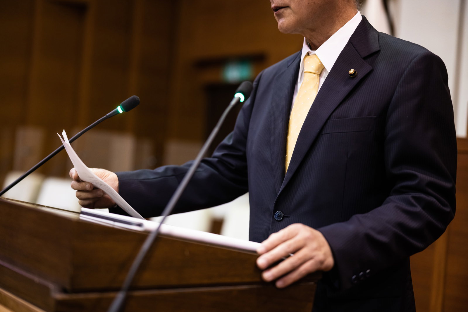 市議会で資料を手に持ちマイクの前で演説する紺色の背広姿の議員