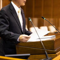 木製演壇で資料を手に発言するダークスーツの男性の写真