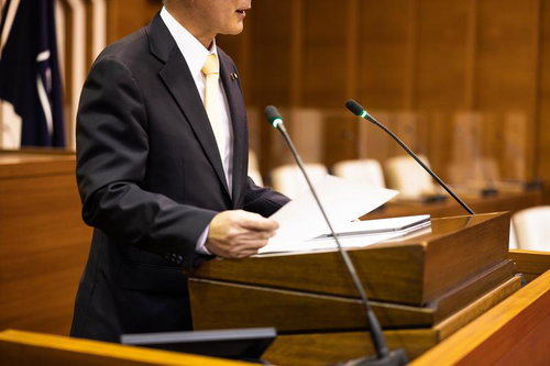 木製演壇で資料を手に発言するダークスーツの男性