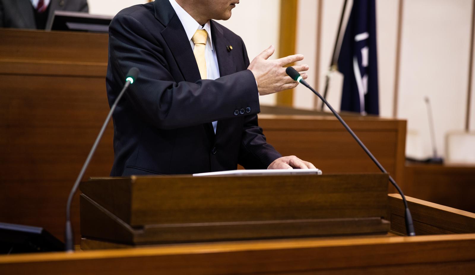 A man in a navy blue suit raising his hand while speaking on stage