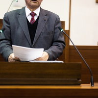 議場の演壇で書類を手に質問する男性議員の議会活動風景の写真