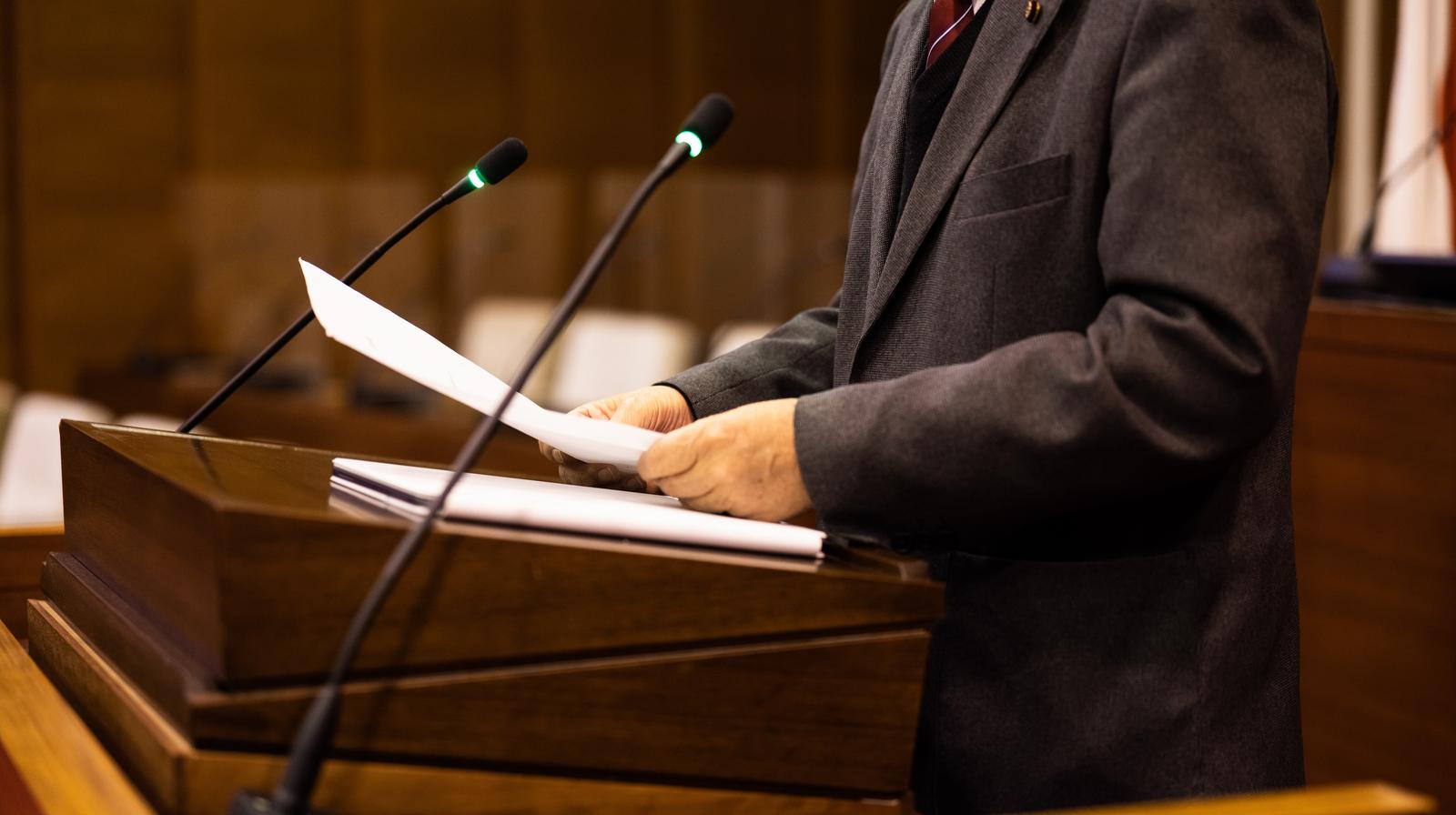 A male councilor in a suit standing at the podium with a microphone and documents in the Tsuyama City Council chamber