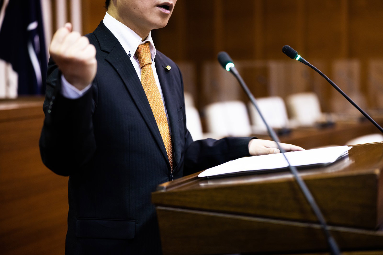 紺のスーツに金色ネクタイの議員がガッツポーズで議会で熱弁する