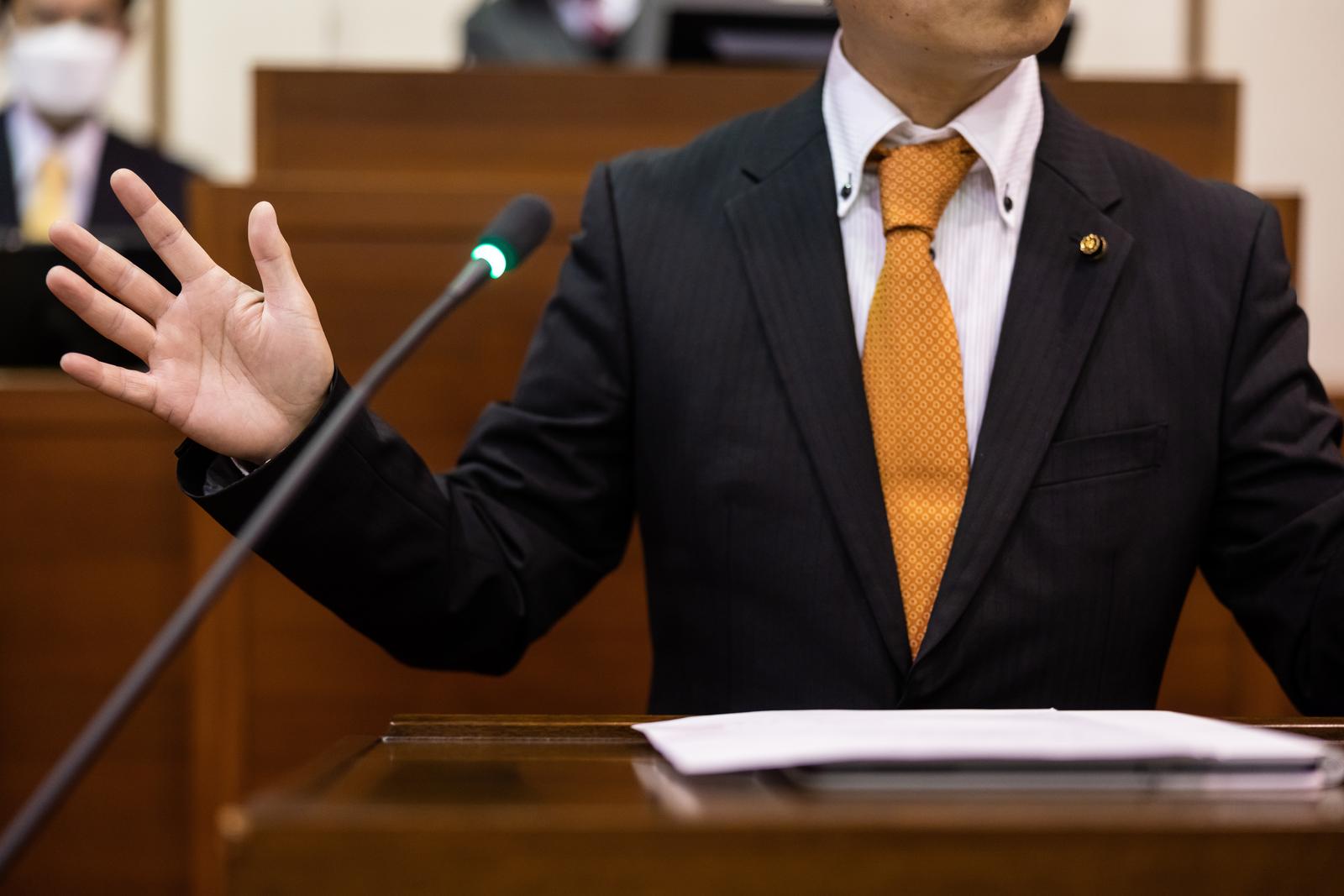 議場で右手を上げながら説明する紺色のスーツ姿の市議会議員