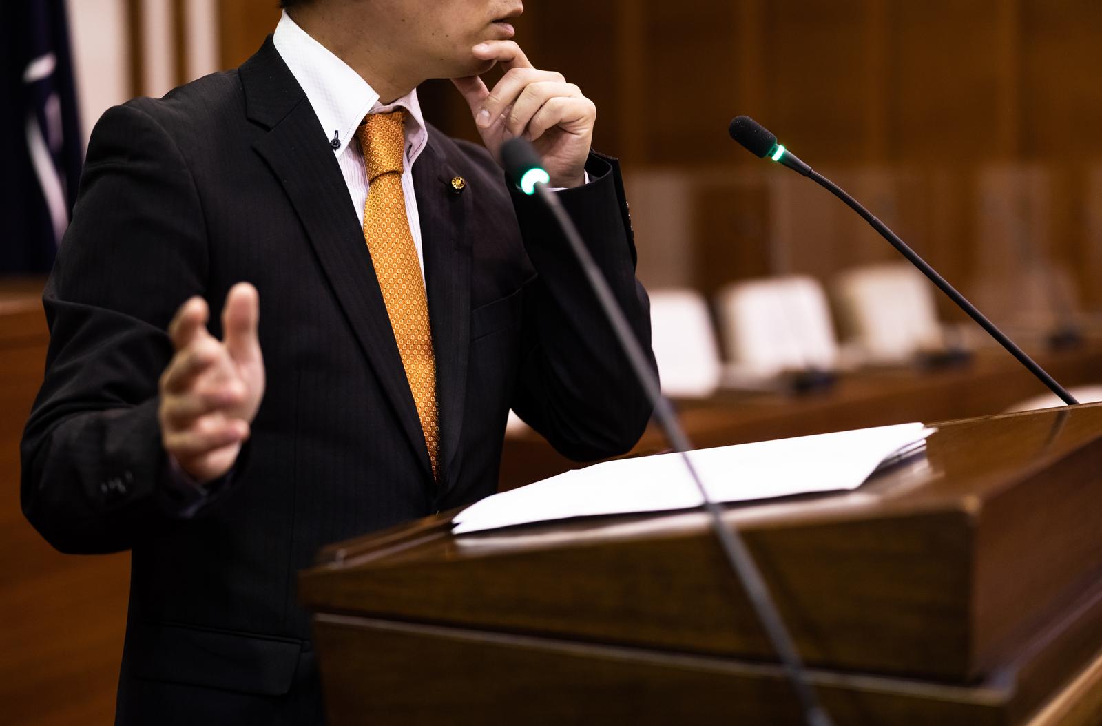 議場で身振りを交えながら発言する黄土色ネクタイの男性議員