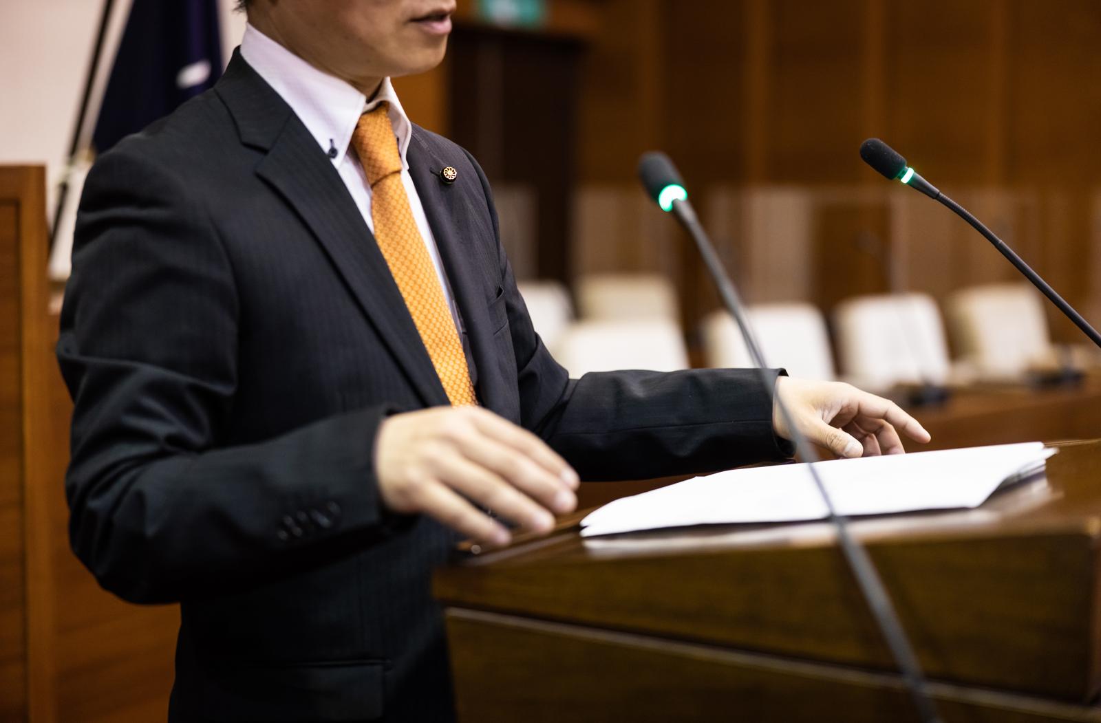 市議会で机の上の書類を見ながら発言する男性議員