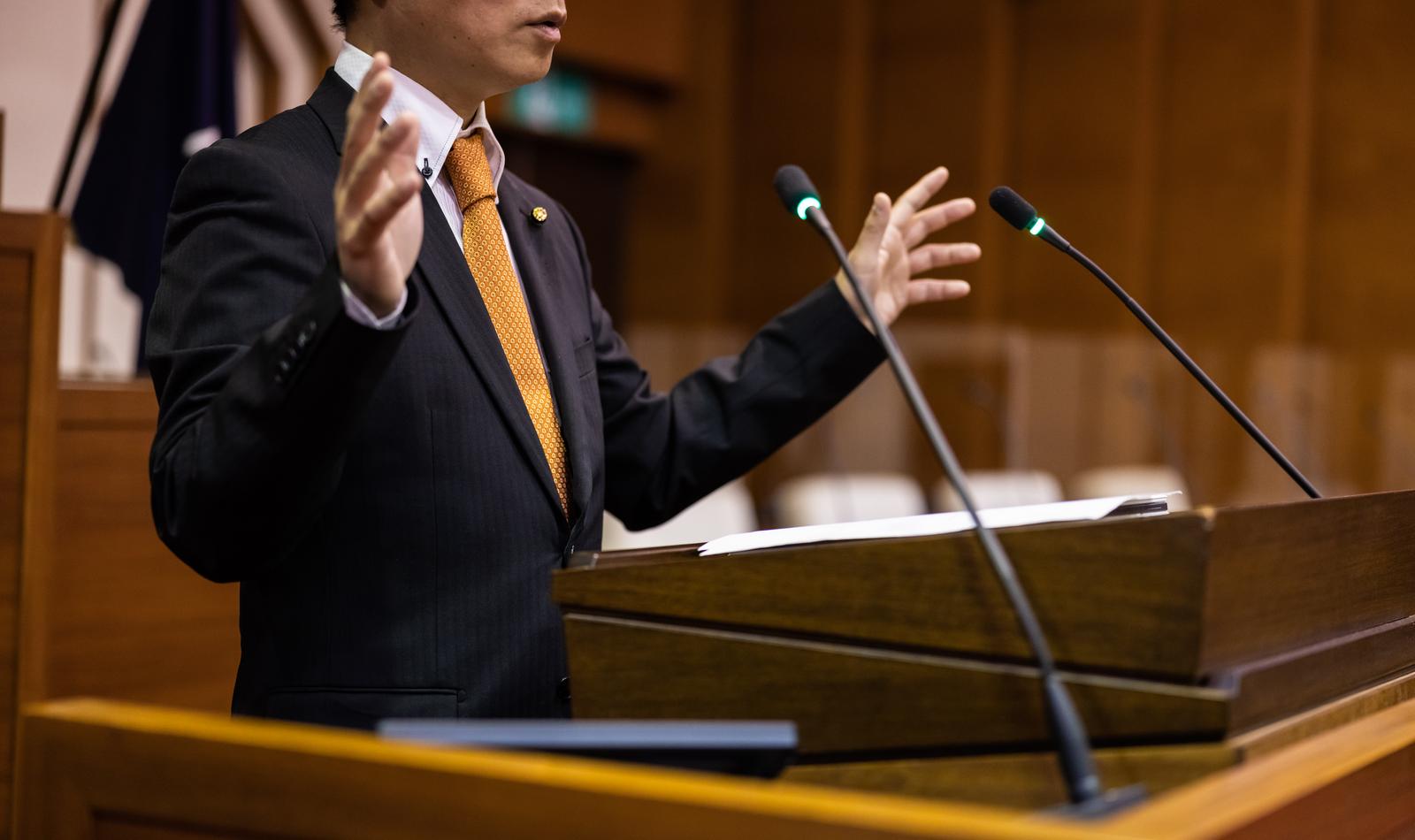 紺色のスーツに金色のネクタイを着けた男性議員が議会の演壇でジェスチャーをしながらスピーチしている