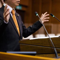 議会の演壇でジェスチャーを交えてスピーチをする市議会議員の写真
