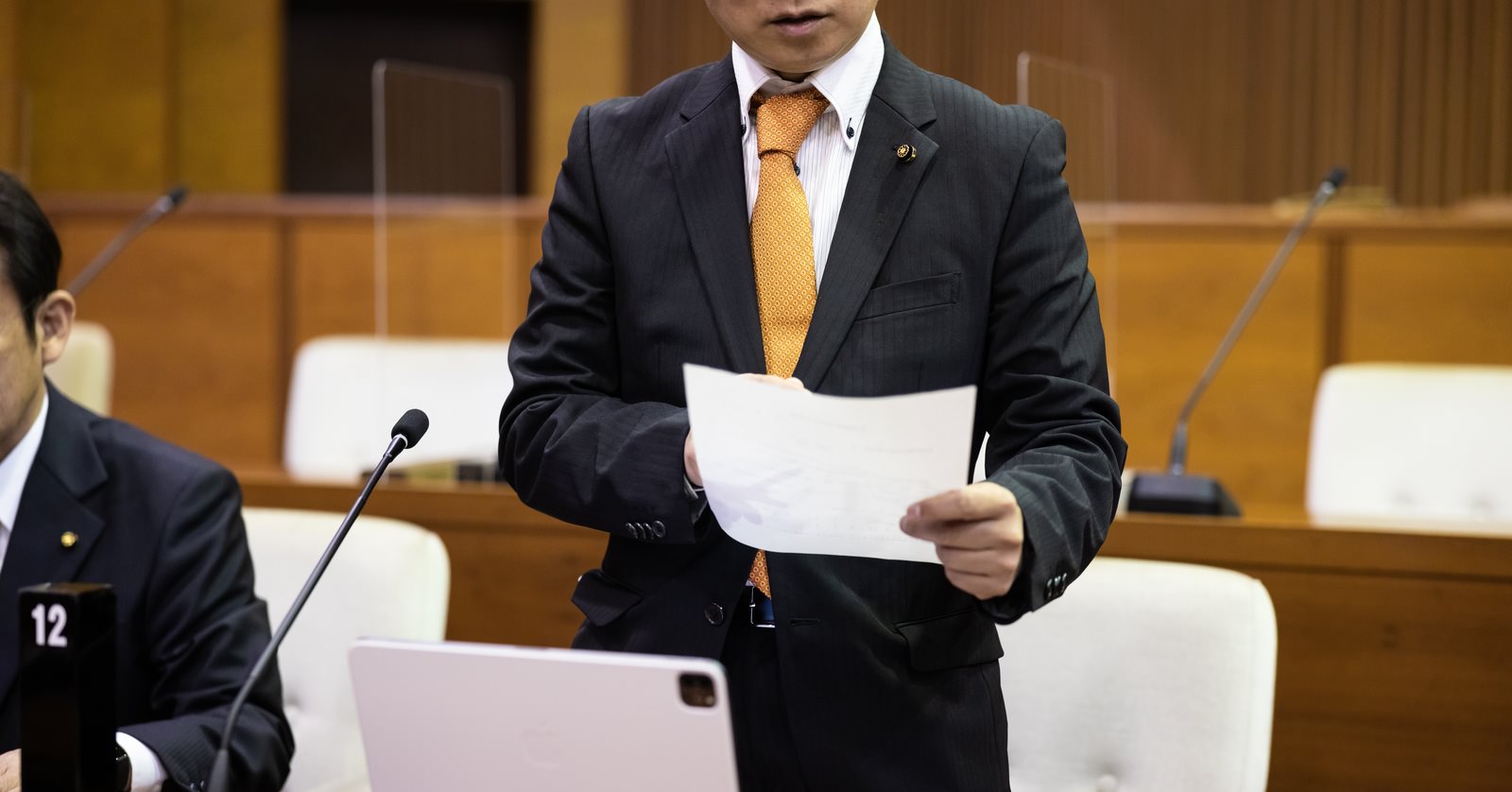 市議会の議場で紙資料とタブレットを使いながら発言する議員