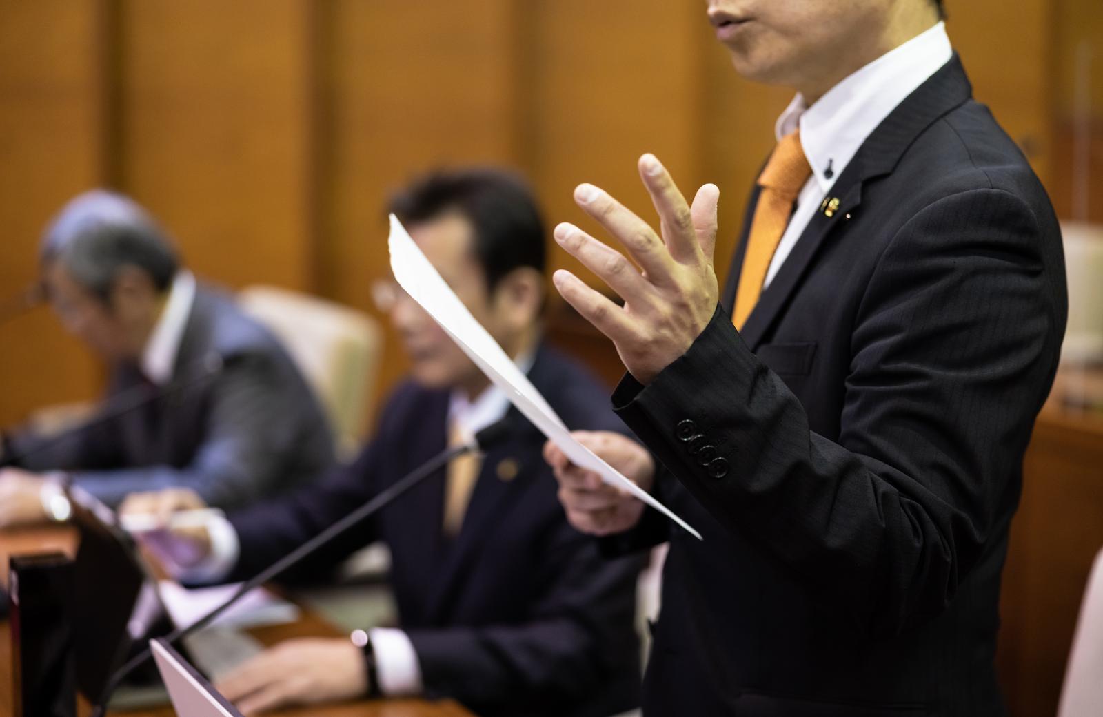 紙の資料を持ちながら議論する黄色いネクタイの議員と議会の風景