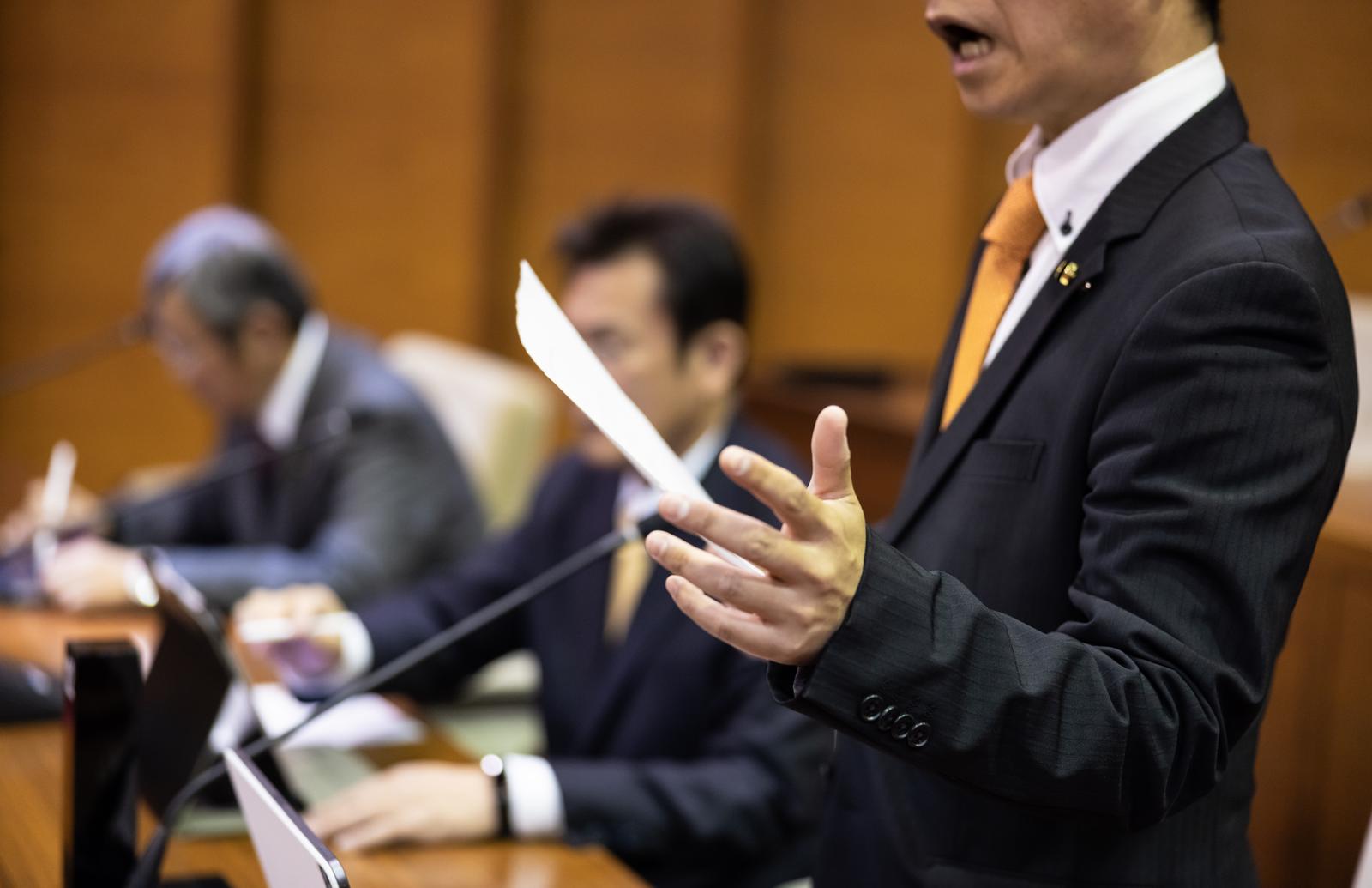 津山市議会の議場で手振りを交えて発言する黄色いネクタイの議員