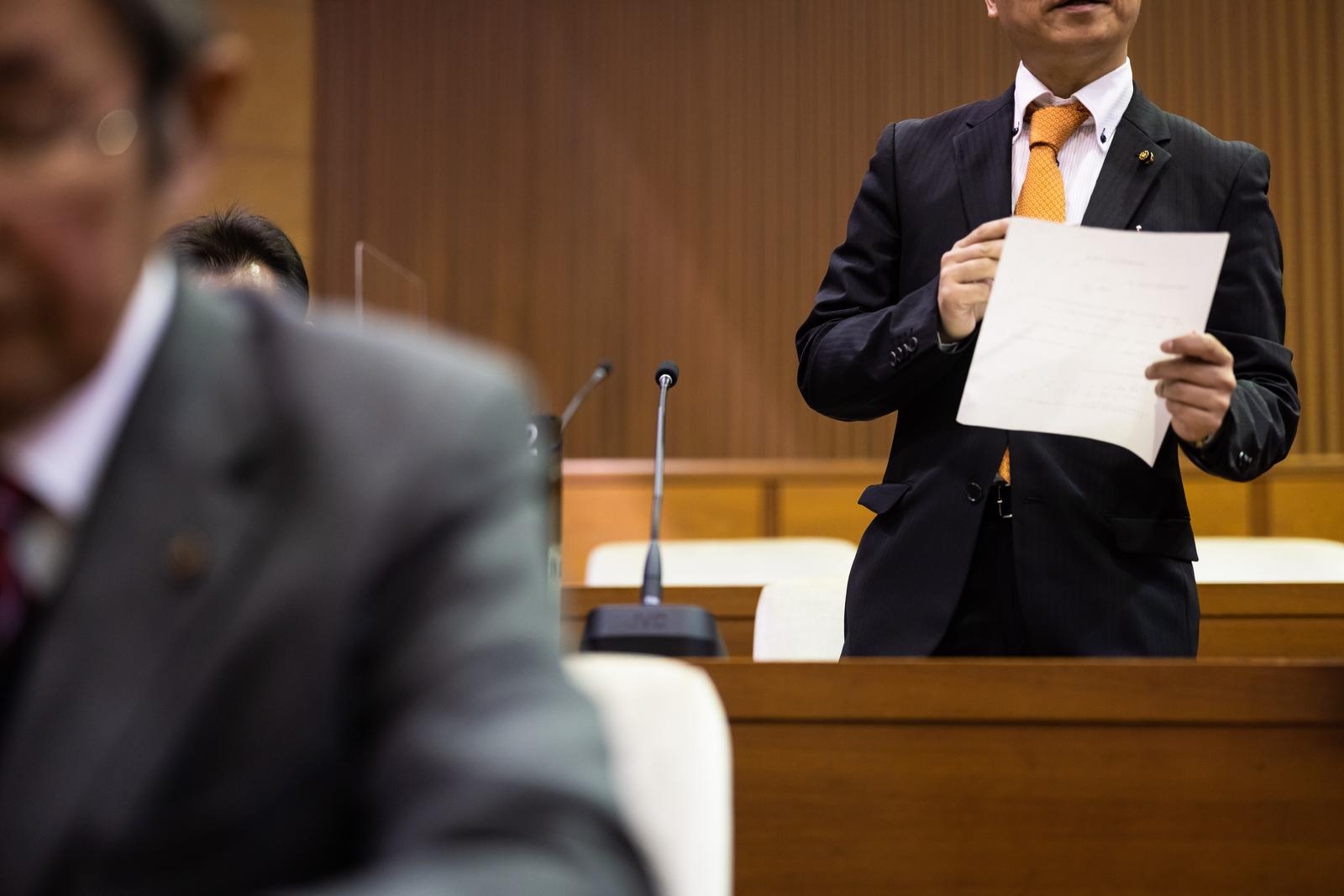議場で白い書類を持ち意見を述べる紺色スーツの男性議員