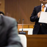 議場で書類を手に意見を述べる紺色スーツの男性議員の写真