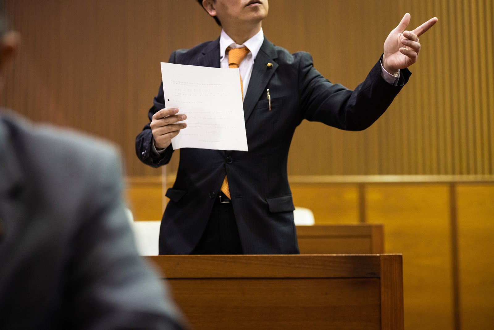 市議会の議場で資料を手に熱く質問する紺色スーツの男性議員