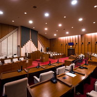 木製パネルと赤い絨毯が特徴の議場、議長席と議員席が並ぶ市議会の写真