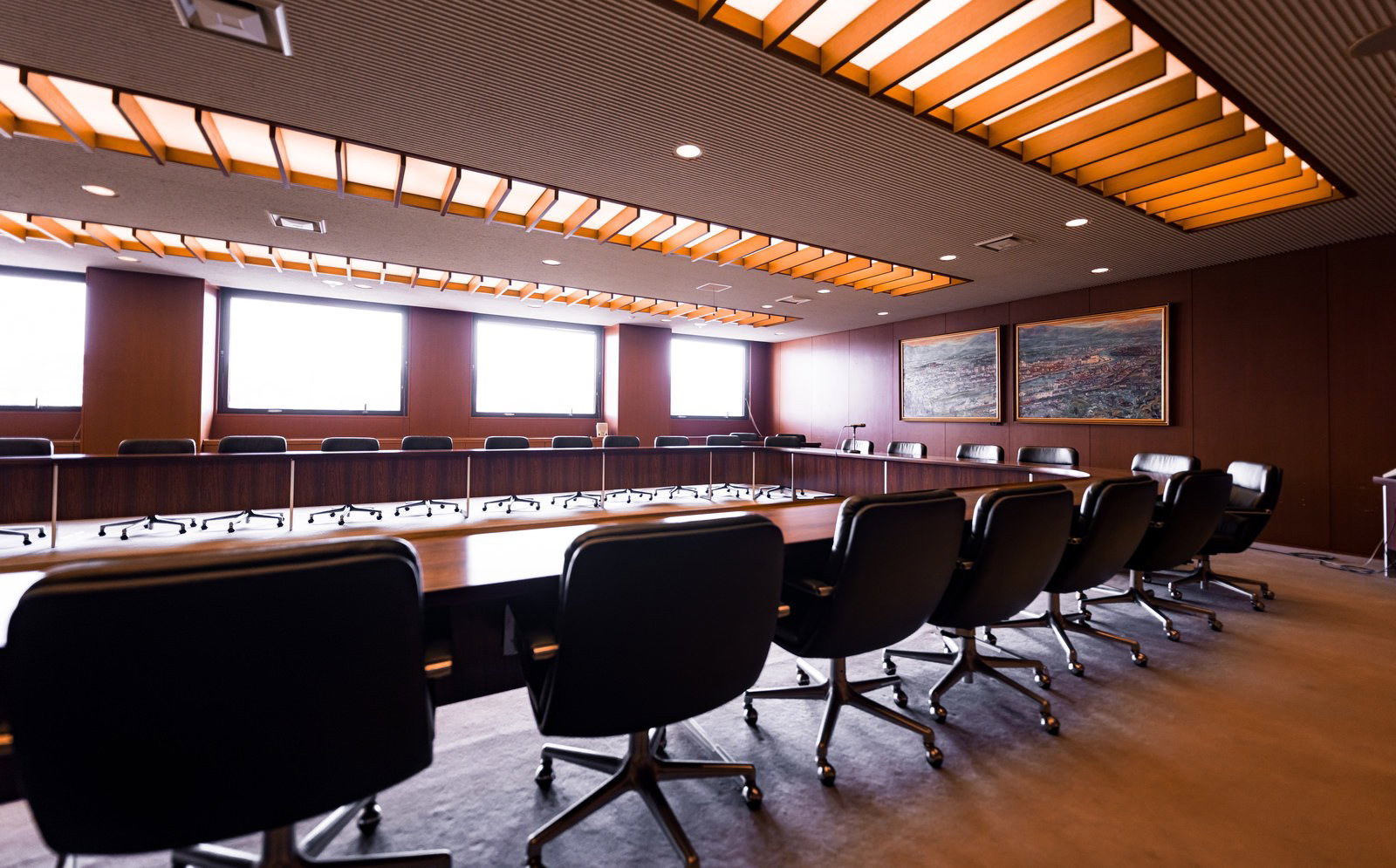 津山市議会全員協議会室。木製テーブルと黒い回転椅子、採光ルーバー天井