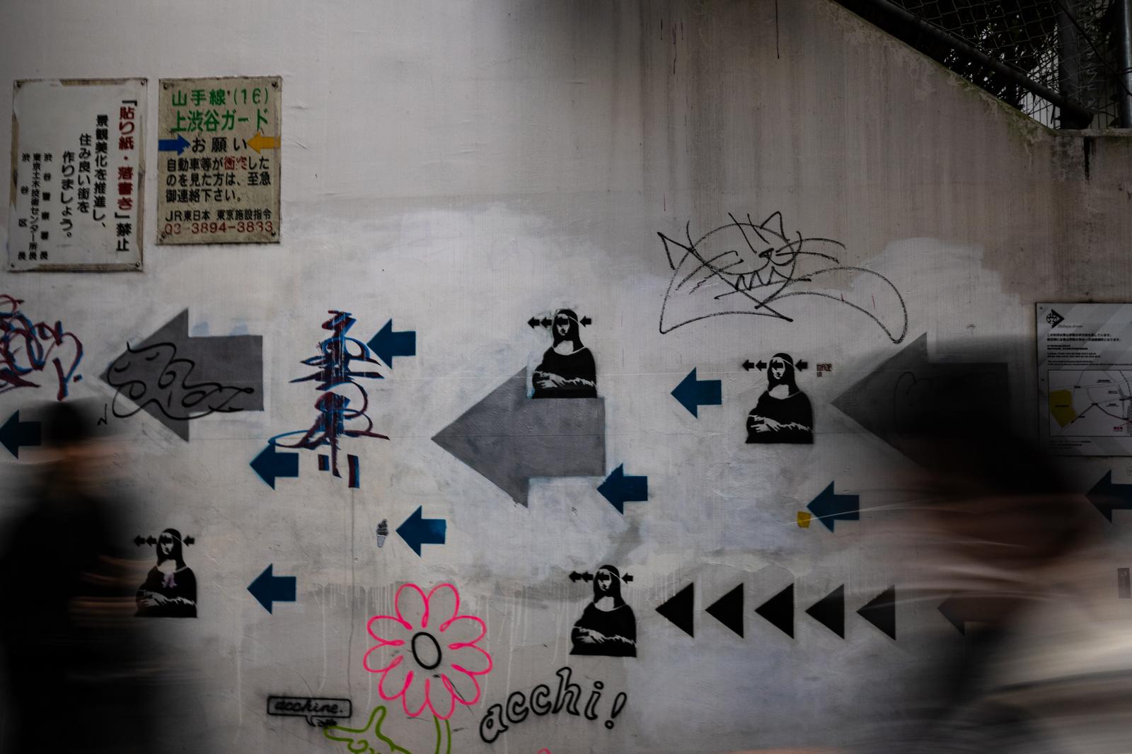 高架下の白い壁に矢印と人物モチーフの壁画が描かれている