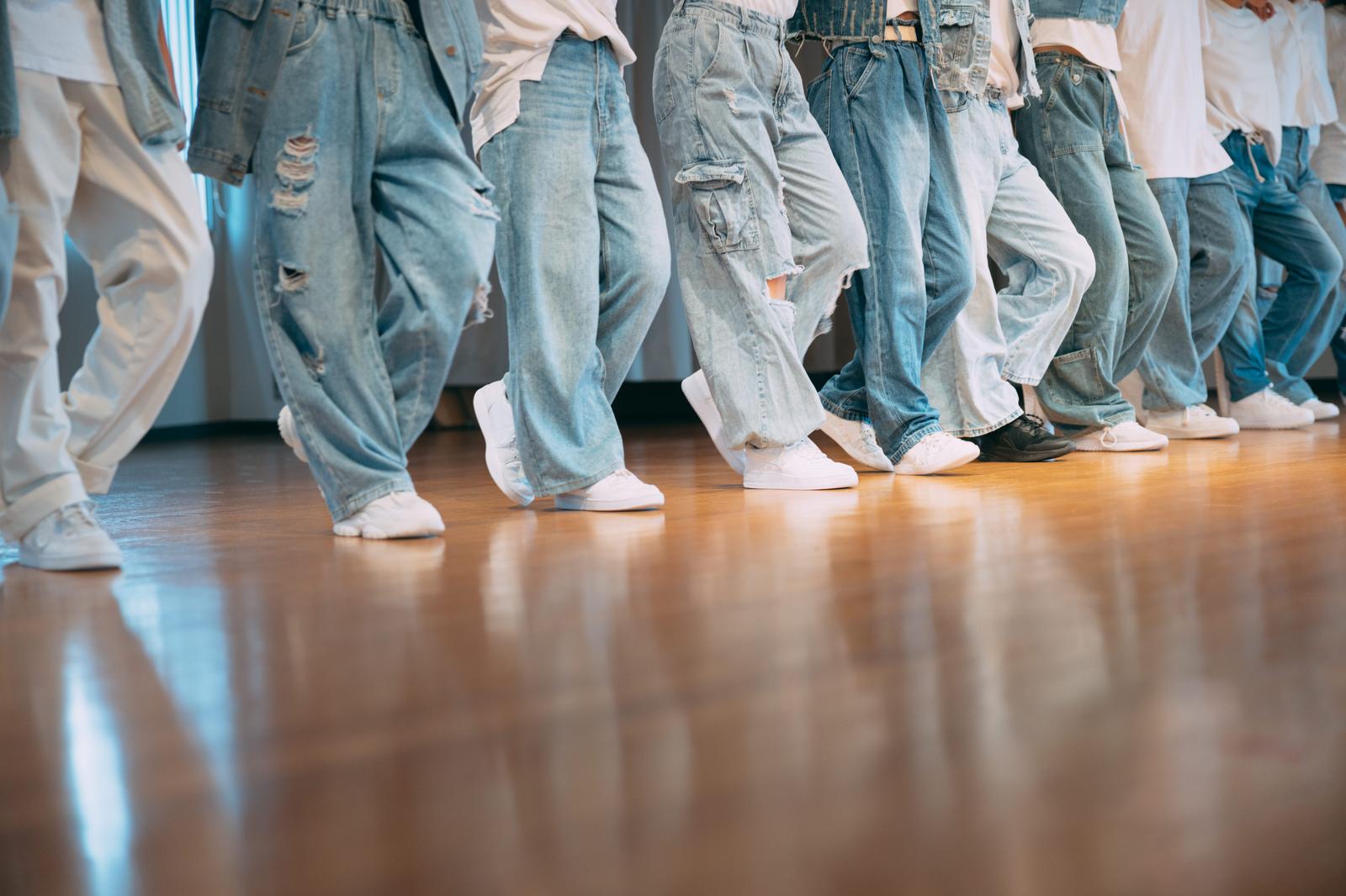 複数人がジーンズ姿で横一列に並び、白いスニーカーでステップを踏むダンスレッスンの足元