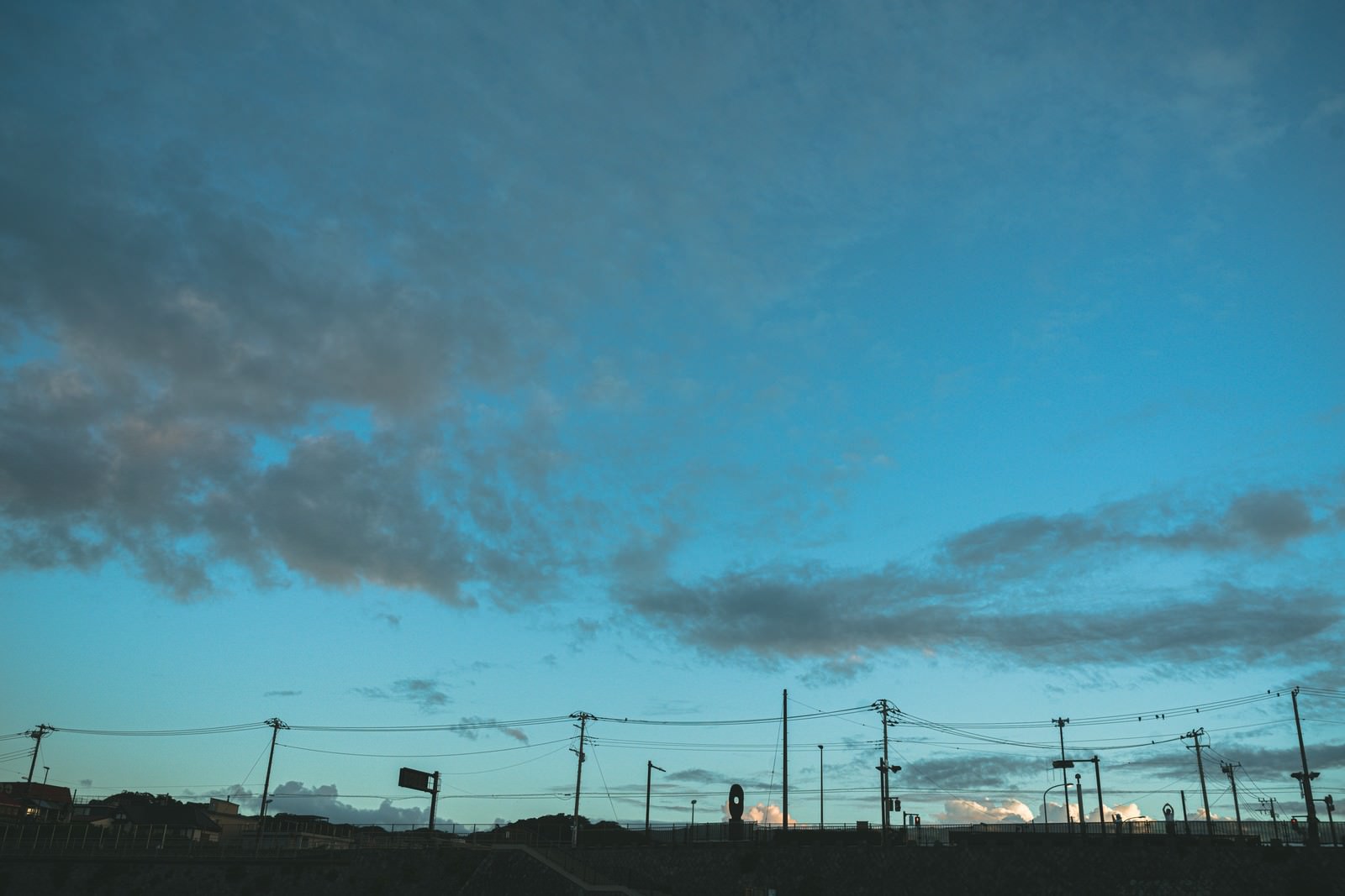 薄雲の浮かぶ青空をバックに、複数の電柱と電線がシルエットで写っている風景