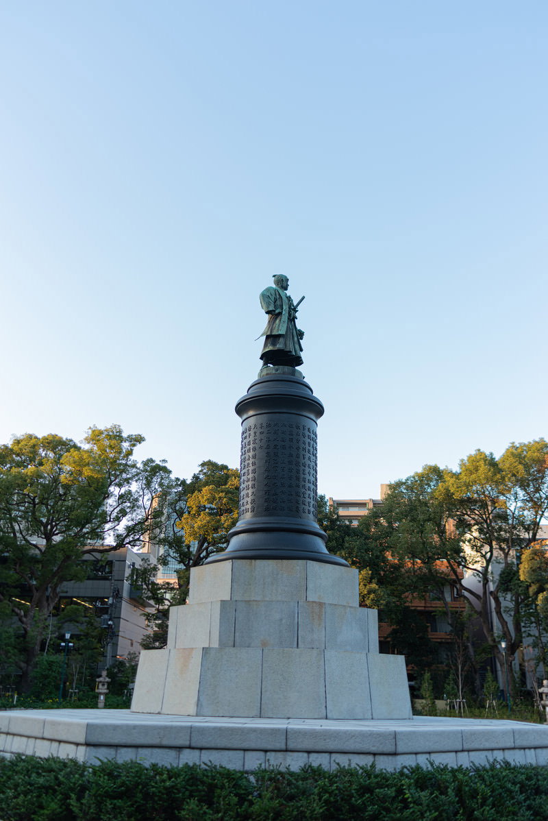 The bronze statue of Okubo Toshimichi standing on a pedestal, with trees around it.