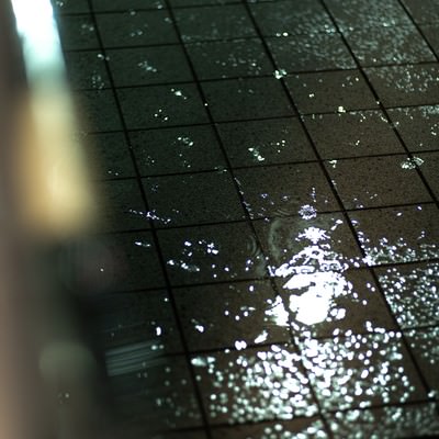 雨降りの水溜りに映る光と水滴、濡れたタイル表面の反射の写真