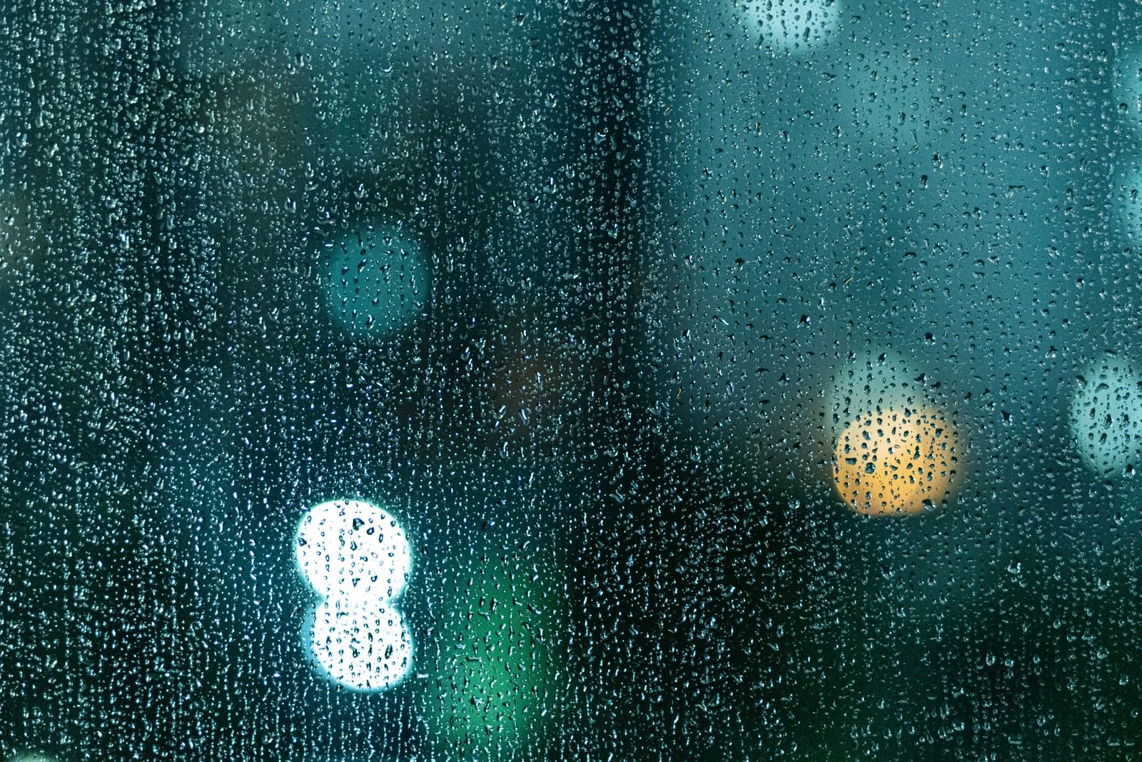 ガラス窓に付着した無数の雨粒と背景にぼんやりと光る暖色の光