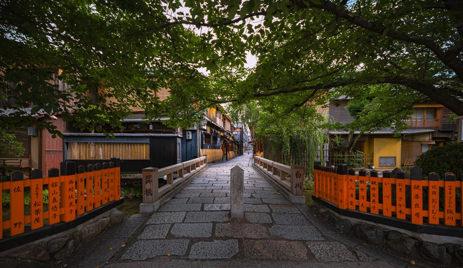 京都祇園白川の朱色の欄干を持つ古い木製の橋、両側に町家が並び樹木に覆われた石畳の路地