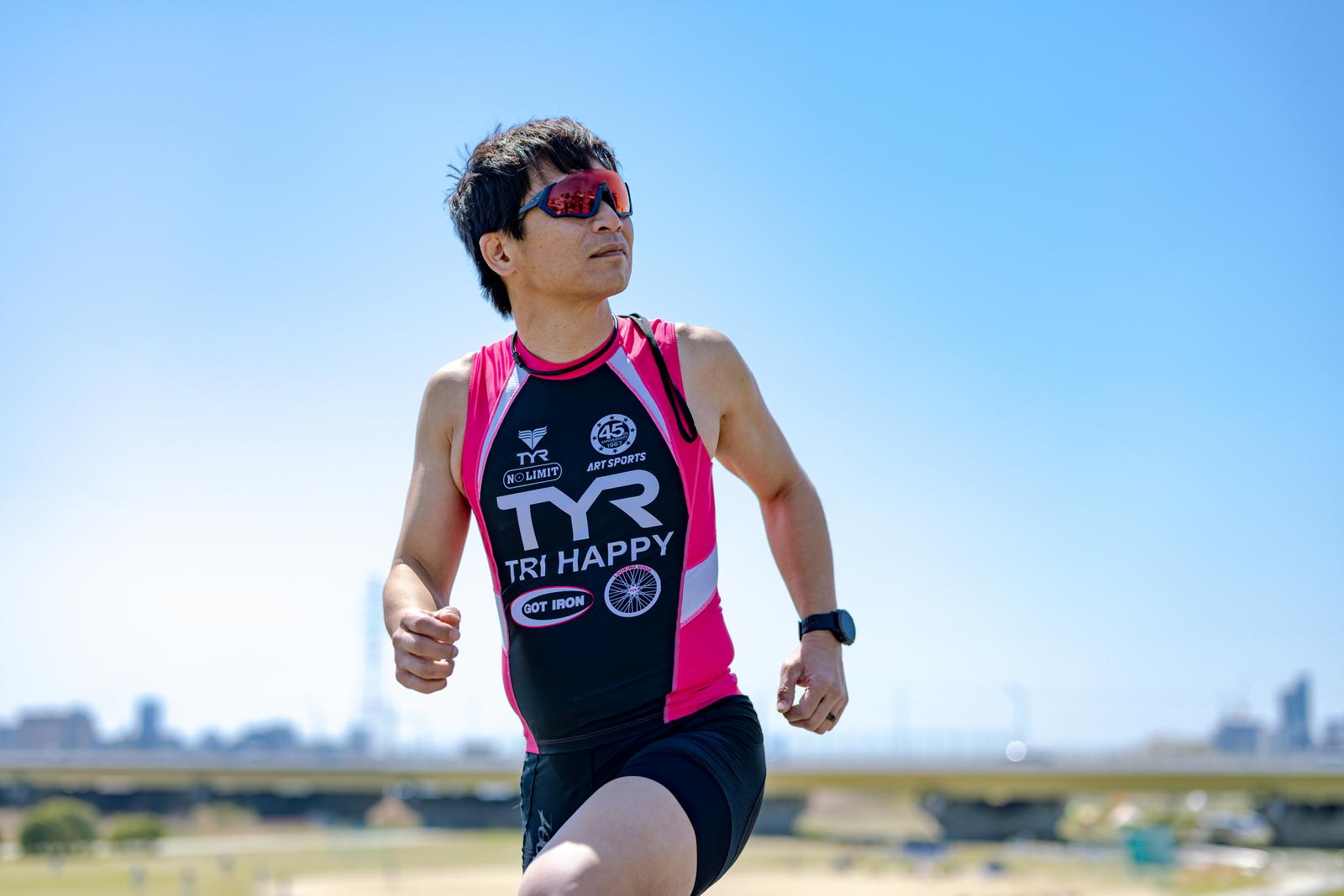青空を背景にサングラスをかけてランニングする男性［モデル：うえせい］