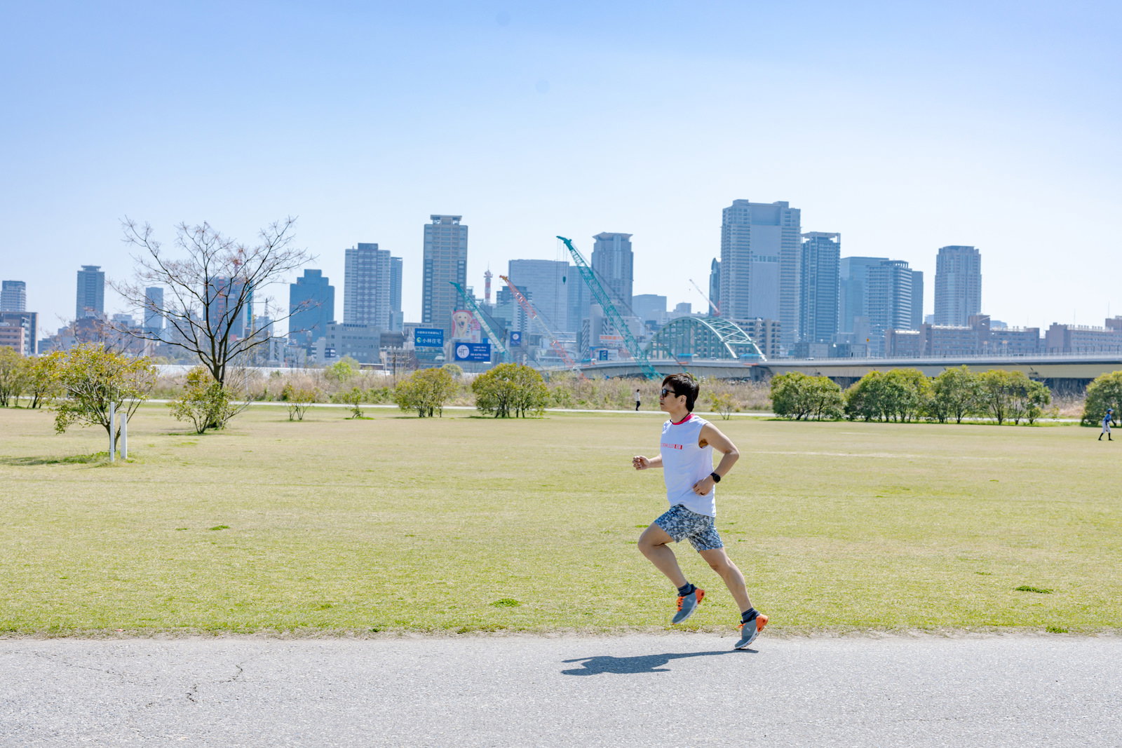 青空と都市景観を背景に芝生エリアでランニングするトライアスリートの男性［モデル：うえせい］