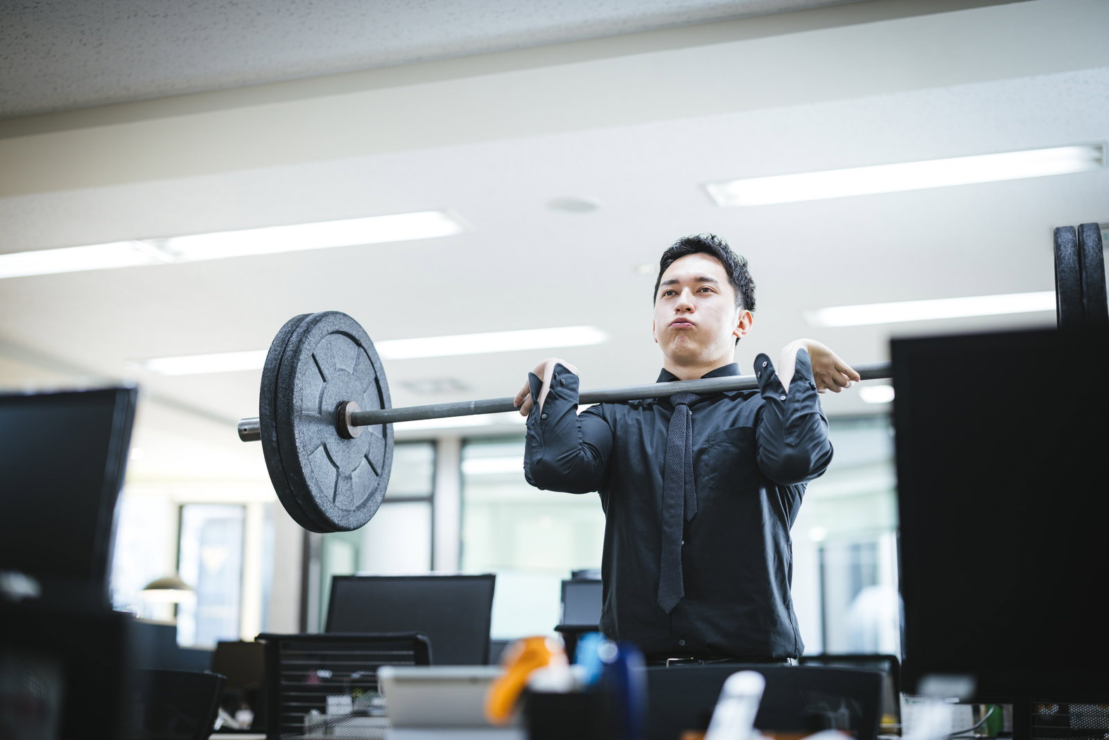 オフィスでバーベルを持ち上げるスーツ姿の男性［モデル：鈴木秀］