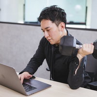 デスクワーク中にダンベルで左腕を鍛えるマッスル経営者の写真