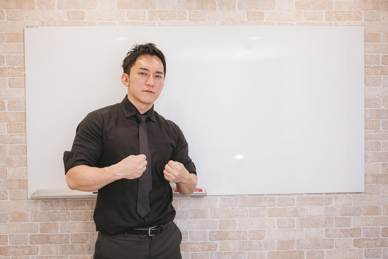 黒いシャツとネクタイを着用した男性がホワイトボードを背に両手を握ってプレゼンしている［モデル：鈴木秀］