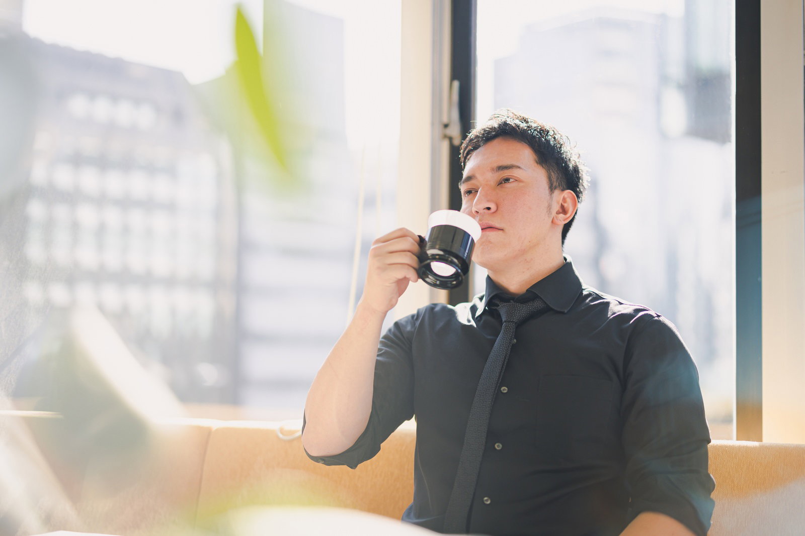 窓際でコーヒーを飲みながら休息する黒シャツの男性経営者［モデル：鈴木秀］
