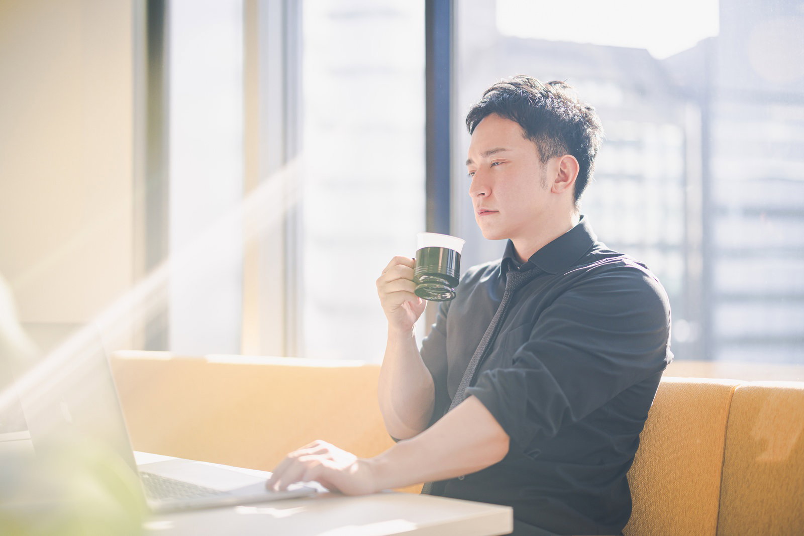 ノートパソコンでデスクワークをしながらコーヒーを飲む男性［モデル：鈴木秀］