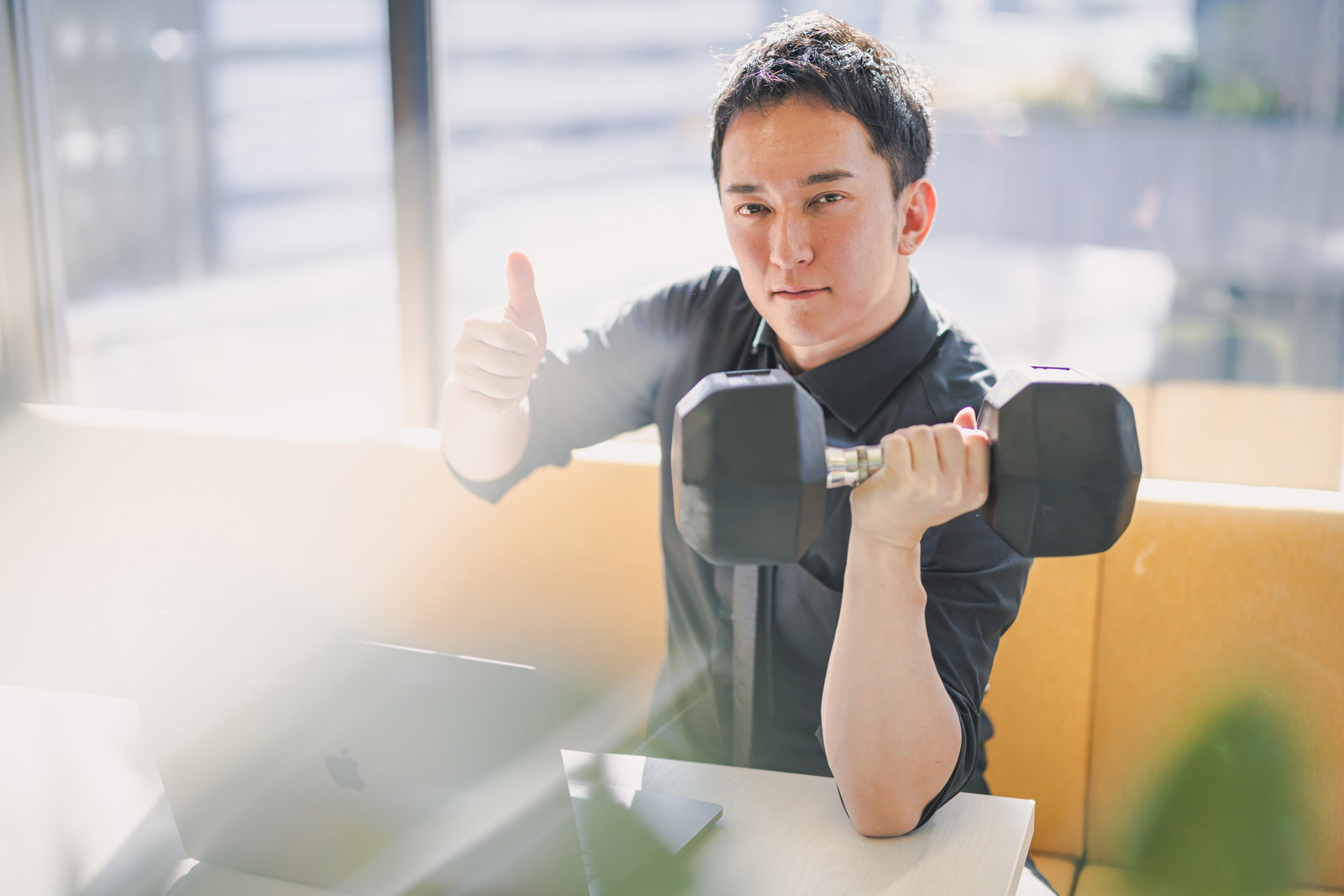 デスクワーク中にダンベルを持ちながらいいねポーズをする男性［モデル：鈴木秀］