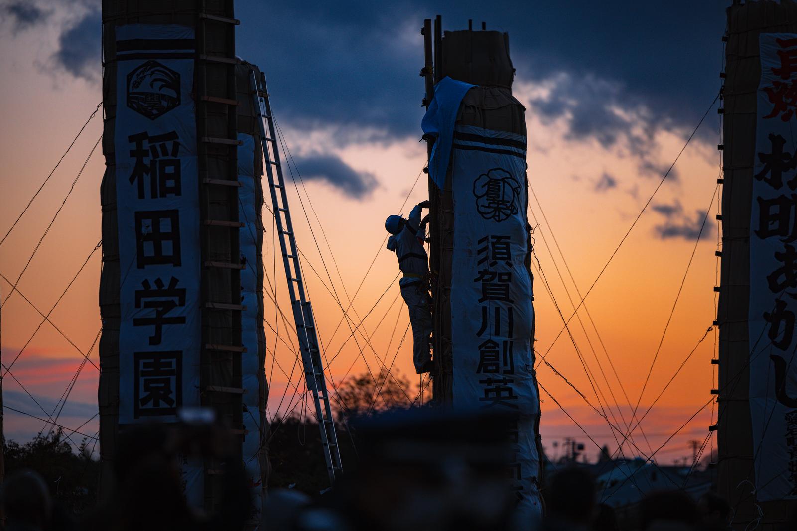 夕焼けを背景に松明の梯子を登るシルエットの作業者