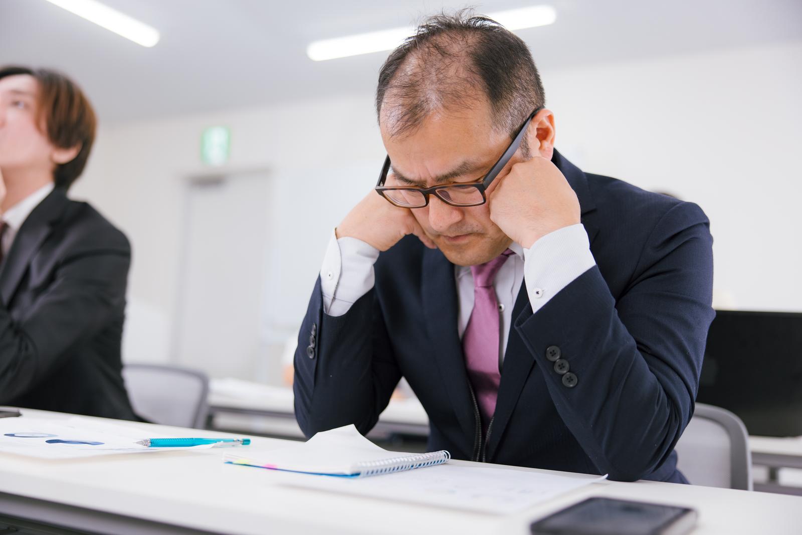会議室で疲労で頭を抱えて俯く眼鏡をかけた男性［モデル：高木桂一］
