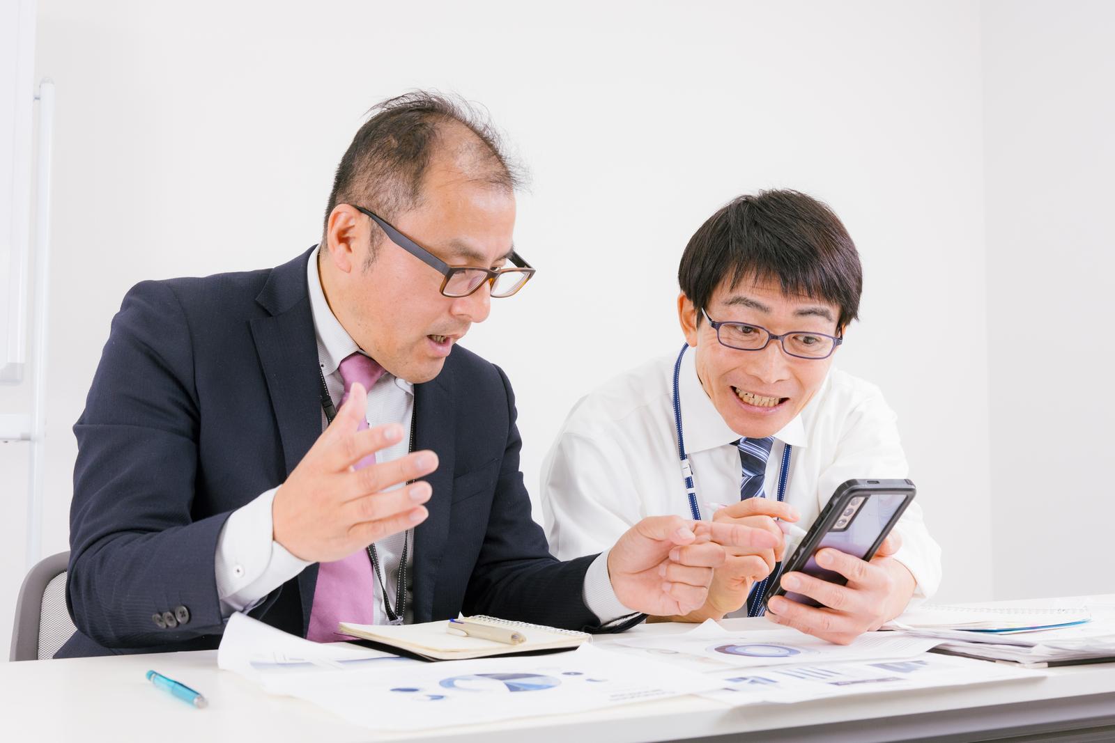 スマートフォンを見ながら打ち合わせするスーツ姿のビジネスマン2人［モデル：高木桂一 ガーミー］