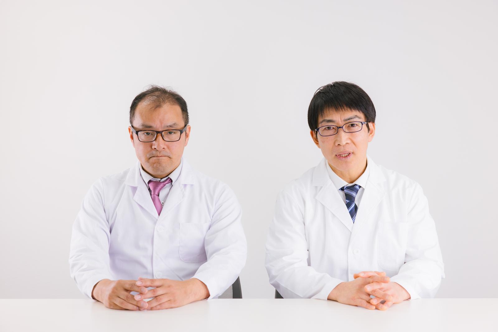 白衣を着た2人の男性医師が真面目な表情でテーブルに向かって座っている［モデル：高木桂一 ガーミー］