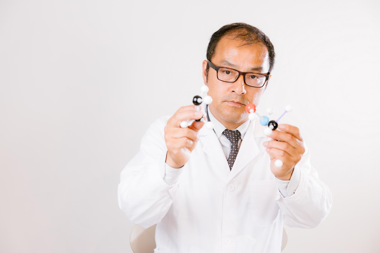 眼鏡をかけた白衣の男性が、カラフルな分子構造模型を両手で持っている［モデル：高木桂一］