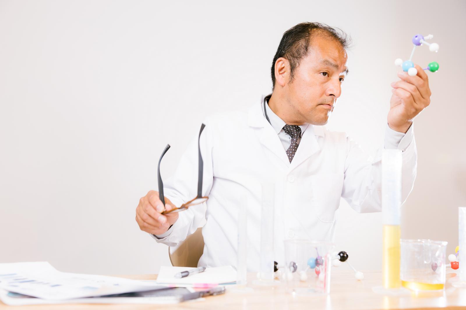 白衣を着た男性科学者がカラフルな分子構造モデルを手に取り観察している［モデル：高木桂一］