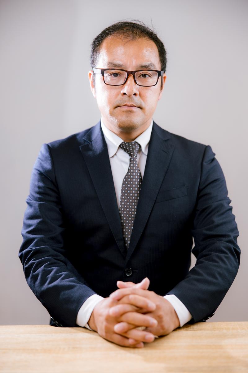 眼鏡をかけたスーツ姿の男性がネクタイ姿で机に向かって座る面接官［モデル：高木桂一］