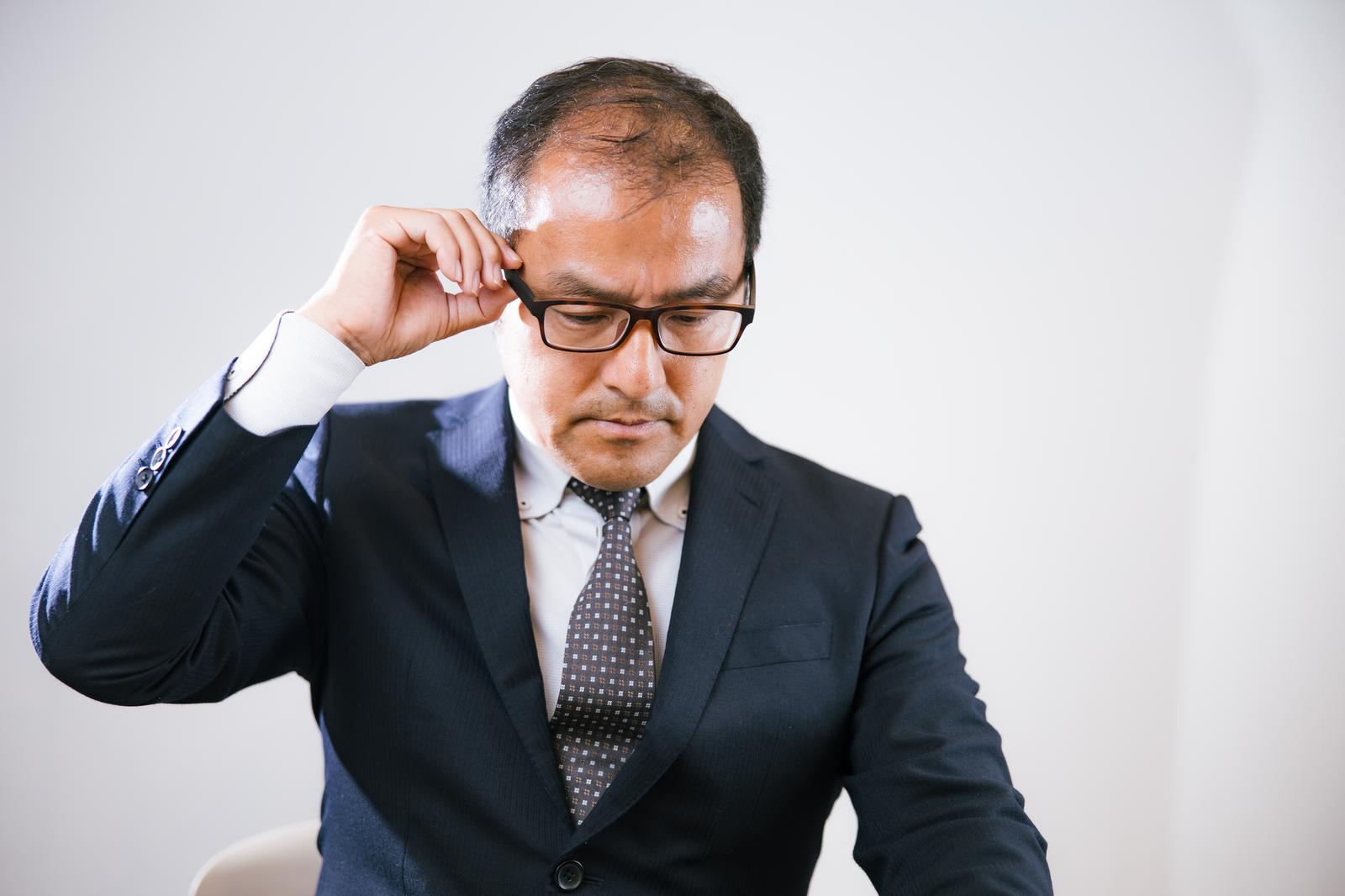 眼鏡を調整しながら下を向くスーツ姿の男性会社員［モデル：高木桂一］