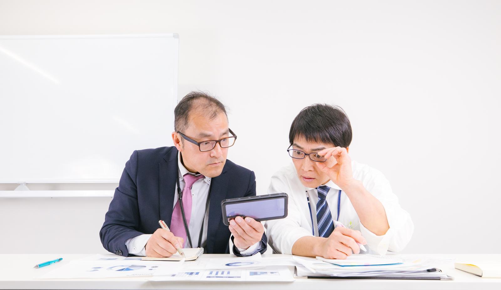 会議室でタブレット端末と資料を確認しながら話し合う2人の眼鏡をかけたビジネスマン［モデル：高木桂一 ガーミー］